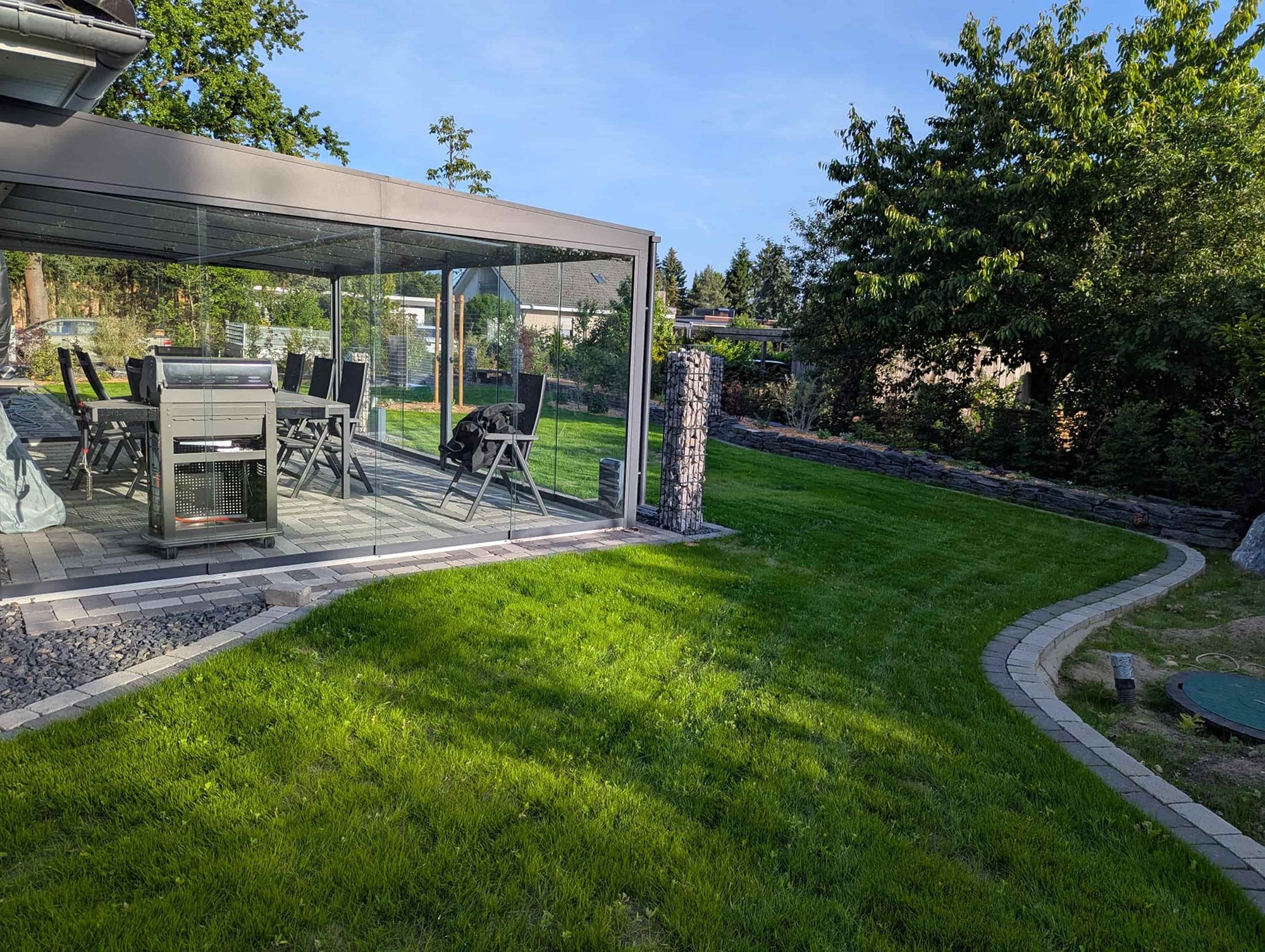 Moderne Terrassenüberdachung mit Glaswänden und Essbereich neben einer geschwungenen Rasenkante in einem sonnigen Garten